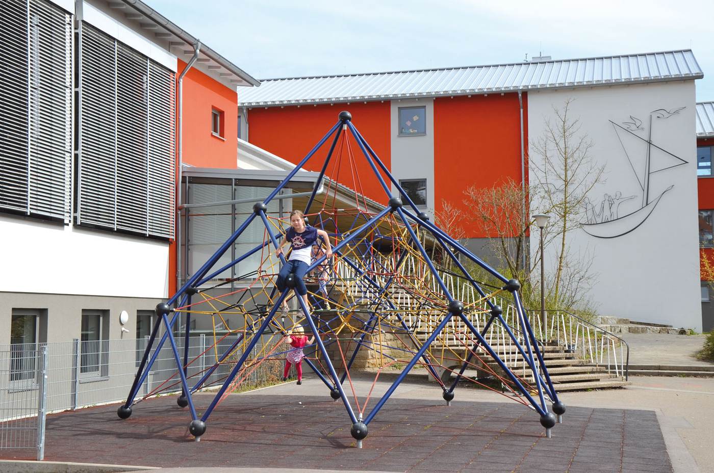Kinder klettern auf der Seilpyramide im Pausenhof vor dem orangefarbenen Schulgebäude